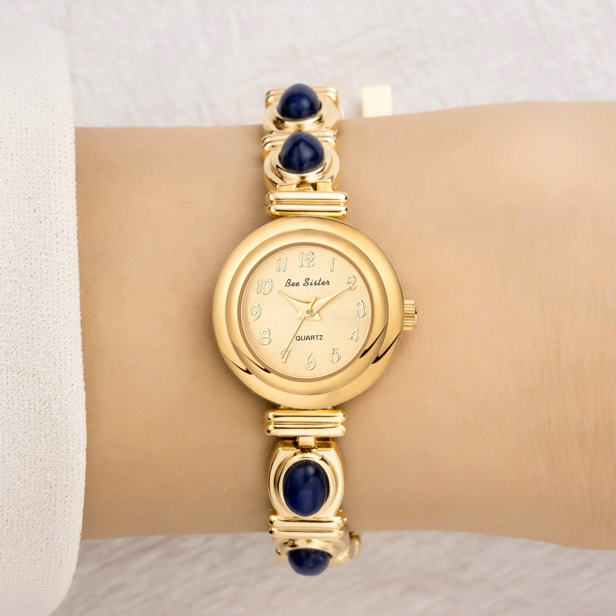 Gold wristwatch with blue gemstones on a wrist against a neutral background